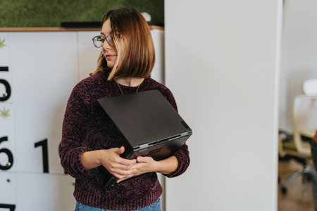 Lovely woman holding a lap top looking aside while leaning on a locker boxes at the office.の写真素材