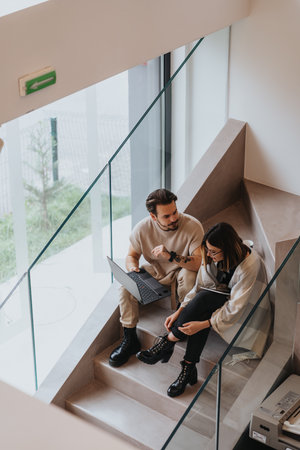 Focused coworkers in a modern office sit on stairs, analyzing statistics and discussing project solutions.の写真素材
