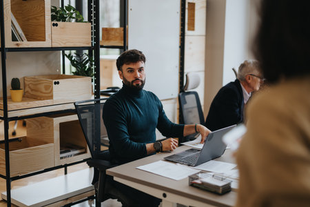 Bearded young man in a turtleneck intensely focusing on his laptop work in a contemporary wooden...