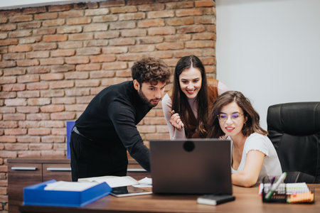 Three professionals collaborating at a laptop in a modern office with a brick wall backdrop, displaying teamwork and engagement.の写真素材