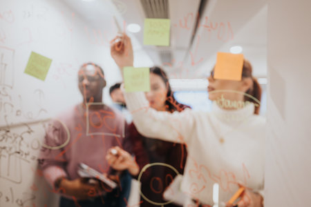 Diverse colleagues brainstorming and strategizing in a pleasant office, using sticky notes on a glass door to create a successful project plan.の写真素材