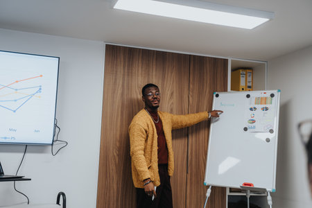 Young man in smart casual outfit pointing at a whiteboard with colorful charts and graphs during a...
