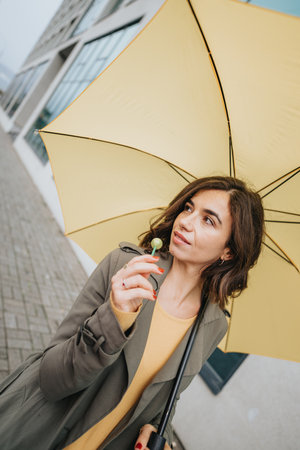 A thoughtful young woman gazes upward while holding a bright yellow umbrella on an overcast day, standing in an urban setting.の写真素材