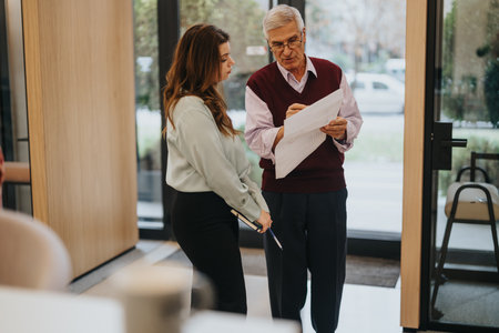 A focused elderly businessman in a smart sweater and a young woman discuss a report. The indoor setting suggests a collaborative atmosphere.の写真素材