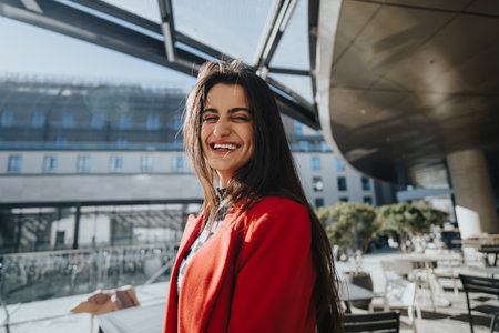 Cheerful young woman in red blazer smiling on sunny city terrace.の写真素材