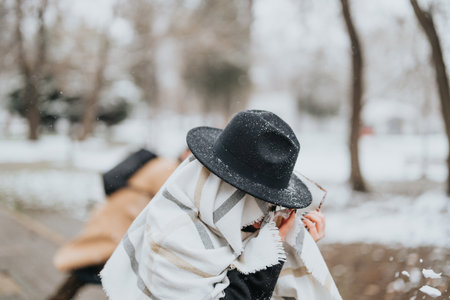 Young professional in stylish hat and scarf enjoying snowy day in park. Girl hiding herself while her boyfriend is throwing snowballs at her.の写真素材