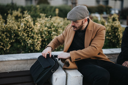 A well-dressed man in a fashionable coat and cap is sitting on a park bench, carefully unpacking his modern leather bag.の写真素材