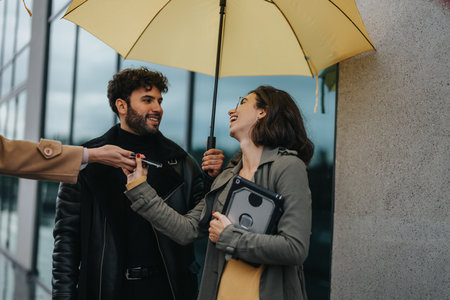 Two smiling friends enjoying a rainy day in the city, sharing a yellow umbrella while laughing and connecting.の写真素材