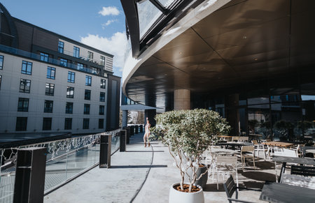 Bright sunlight bathes a contemporary outdoor terrace with stylish furniture and a woman enjoys a solitary walk amidst the urban setting.の写真素材