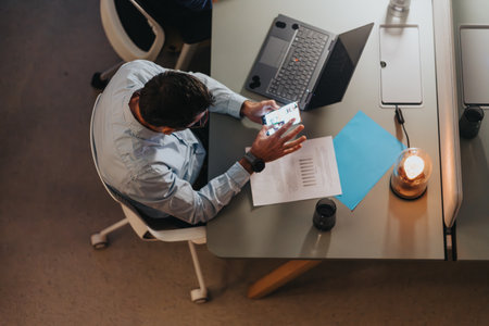 Professional senior man checking smart phone at modern workspace with laptop and documents.
