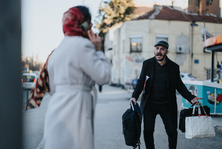 In an urban setting, a young businessman holding multiple bags confronts his distracted girlfriend who is engaged in a phone conversation.の写真素材