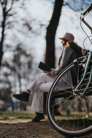 Stylish young woman relaxes on a bench in an urban park, with her vintage bike beside her, enjoying a peaceful sunny day.の写真素材