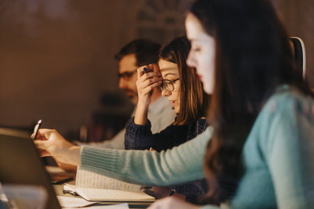 Three focused colleagues working together on a project in a dimly lit office space late at night,...