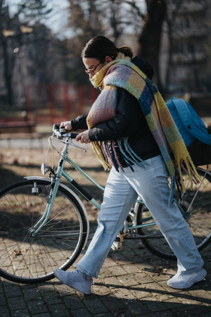 Lifestyle image of a young woman walking alongside her bike on a paved path in an urban park,...