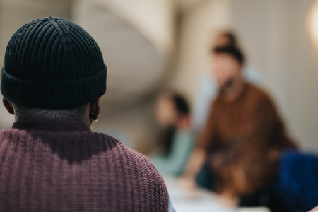 A man in a beanie observes from afar, out-of-focus individuals meet in a softly lit environment,...