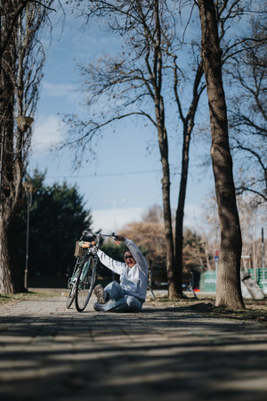 Joyful individual in casual wear laughing and holding a bicycle overhead while sitting on a park path with trees in the background.の写真素材