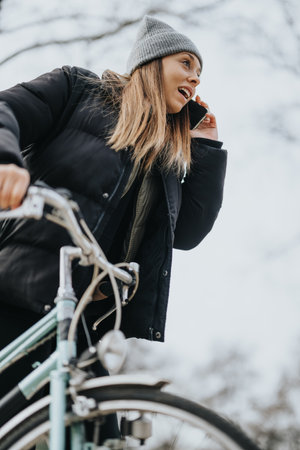 A young woman engaged in a phone conversation rides her bicycle during winter, wrapped up in a coat and beanie against the chilly weather.の写真素材