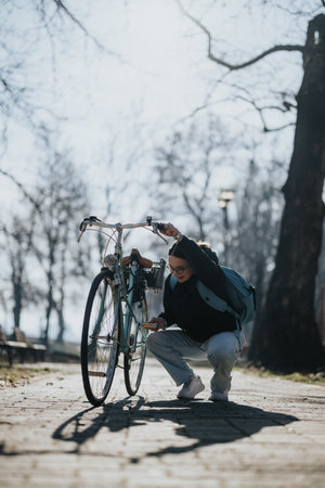 A woman with a backpack pauses during her bike ride to check something on her bicycle on a sunny, tree-lined path.の写真素材