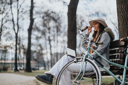 Fashionable young lady in a chic hat sitting on a park bench next to her vintage bicycle, using a laptop on a sunny day.の写真素材