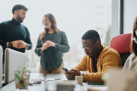 An engaged African American professional checks his smart phone amidst a casual business meeting with colleagues discussing in the backdrop.の写真素材
