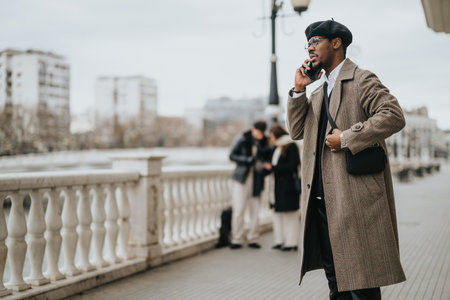 Elegant man in a herringbone coat having a phone conversation while walking on a city bridge, with other pedestrians in the background.の写真素材