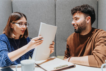 A focused female colleagues presents her ideas from a notebook to a smiling male colleague at a coffee shop, illustrating a casual productive meeting.の写真素材