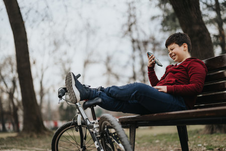 Young boy relaxing on a park bench with a smart phone and bicycleの写真素材