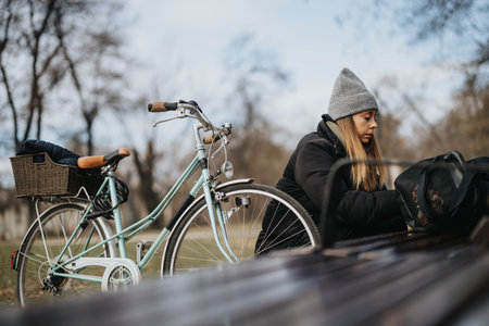 Young female in a wool hat and jacket taking a break on a bench with her stylish city bike beside her on a chilly autumn day in the park.の写真素材