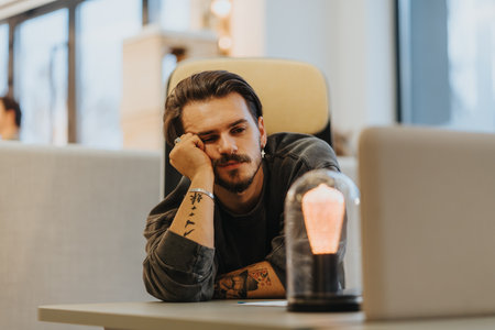Pensive tattooed man with hand on chin sitting at a desk with laptop and light bulb.