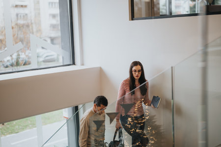 A young professional woman climbs the stairs in a modern office setting, tablet in hand, as a colleague descends with a briefcase.の写真素材