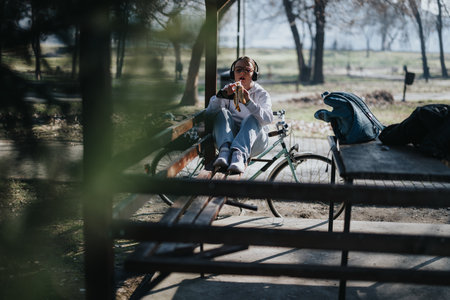 A relaxed young adult female listens to music on her headphones, casually seated on a park bench alongside her bicycle.の写真素材
