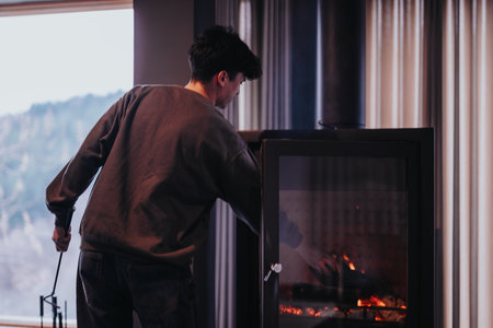 Young male person putting woods at the burning stove, taking care of the fire.の写真素材