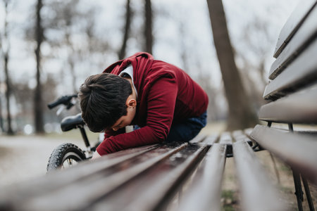 Young boy laying his head down on park bench with bicycle behind himの写真素材