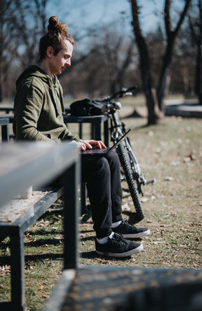 Young adult man with a man bun using laptop in a park with his bike beside himの写真素材