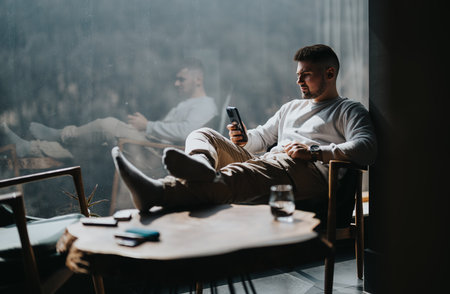 Relaxed man using smart phone in a modern, sunlit room recharging from daily stresses
