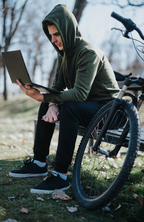 Young man balancing work and leisure by using a laptop in a park, seated next to his bicycleの写真素材