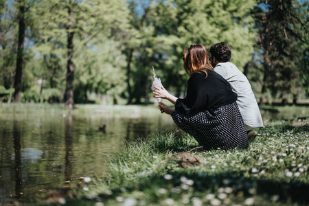 A young couple sits back-to-back by a pond in a lush urban park, displaying a moment of relaxation...
