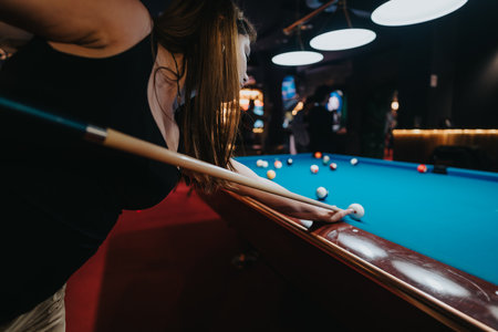 Focused young woman lining up her shot at a billiard game, surrounded by friends in a dynamic, well-lit pool hall.の写真素材
