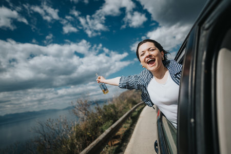 Joyful woman celebrating with beverage while hanging out of car window on scenic road tripの写真素材