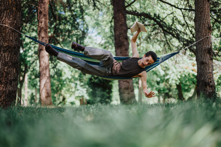 Young man reading and relaxing in a hammock, nearly falling in a lush green parkの写真素材