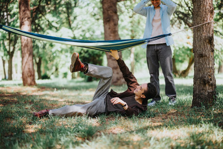 Two young men enjoying friendship in a sunny urban park, one playfully falling from a hammock