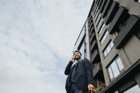 Smiling businessman talking on mobile phone walking outside modern buildingの写真素材