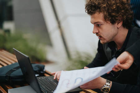 Young man working on a laptop while reviewing documents outdoorsの写真素材