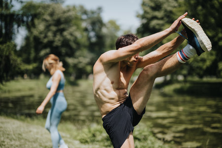 Fit man and woman performing outdoor warm-up exercises by a lake on a sunny dayの写真素材