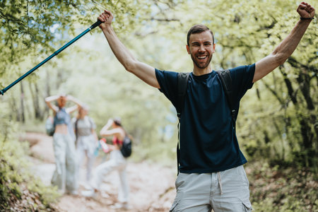 Male hiker standing and celebrating in a lush green forest with people in the background. He is holding a hiking pole in his hand.の写真素材