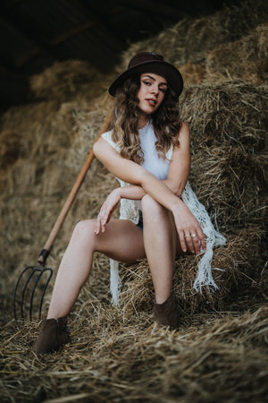 Young woman in rustic setting with hay bales and pitchfork, wearing hat and casual outfitの写真素材