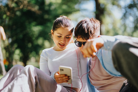 Two teens enjoy music and social media on a smart phone in a sunny parkの写真素材