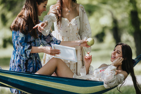 Three joyful friends sharing a moment in a sunny park, relaxing on a hammock with smiles and a bookの写真素材