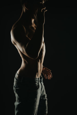 Silhouette of a muscular man with defined abs and jeans in a dimly lit studio