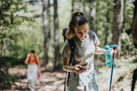 Young woman hiking in a forest checking her phone while holding trekking pole and water bottle